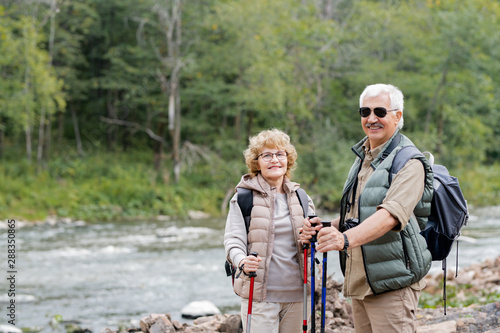 Active aged spouses with backpacks and trekking sticks enjoying summer trip