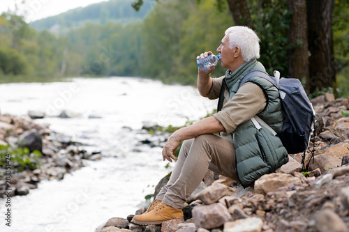 Tired mature backpacker drinking water from plastic bottle on river bank
