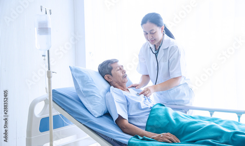 Asian woman professional doctor with notepad smiling, visiting, talking, and diagnosing the old man patient lying in patient’s bed at hospital ward