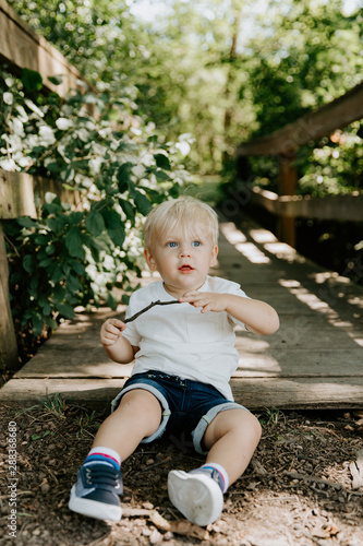 Cute Little Blond Haired Toddler Boy Kid Child Sitting and Laughing in Front of Wooden Bridge Over a Creek at the Outdoor Park in the Forest on a Sunny Pleasant and Happy Summer Day