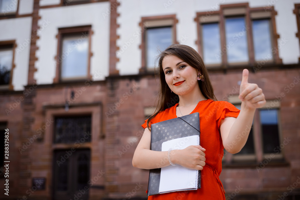 Smiling successful young student showing thumb up against college or university building Smiling successful young student showing thumb up against college or university building