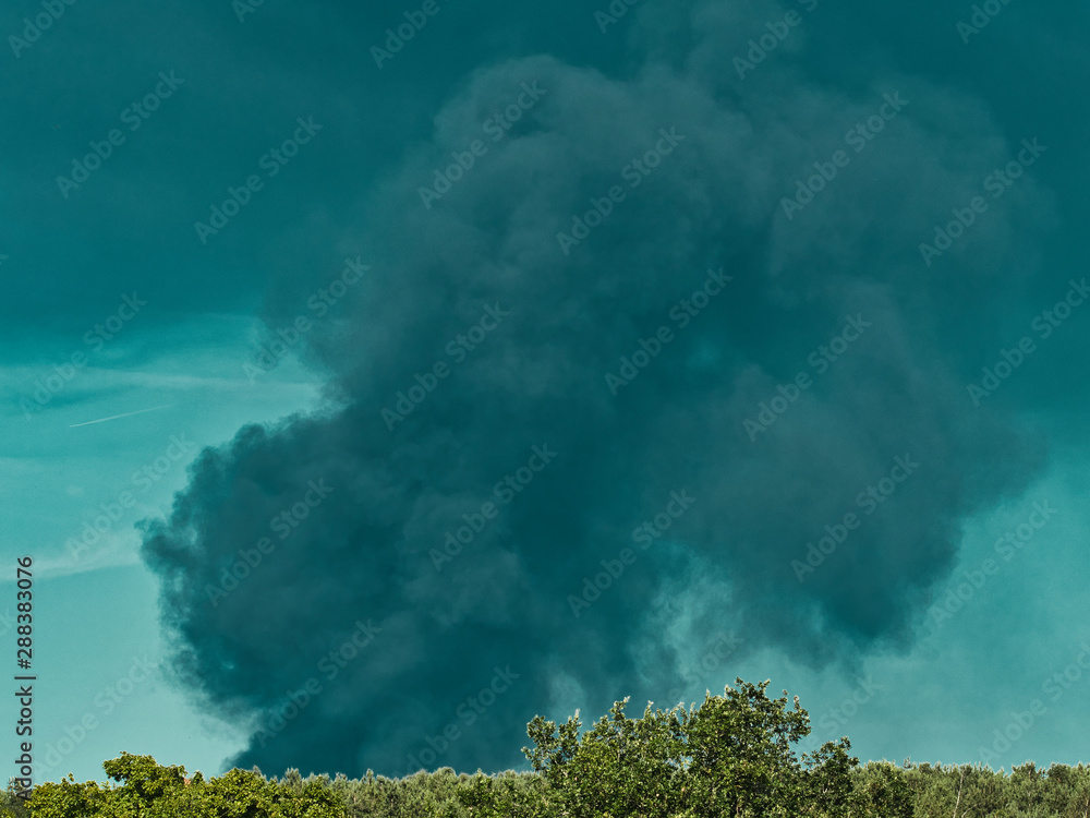 Fototapeta premium A Burning plastic recycling place causes a big pile of dark, black thick smoke on a sky blue summer day. Danger, sorrow. Terrorist attack or explosion.