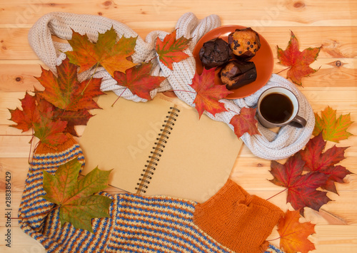 Empty notebook, mug of hot coffee, autumn maple leaves, muffins and warm homemade sweater on wooden surface.