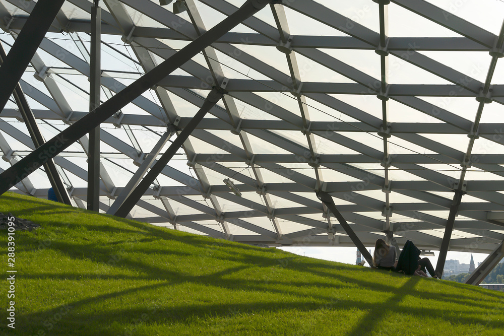 Fototapeta premium Modern ceiling with glass elements, metal mesh with green grass. Blue sky and green landscape. Zaryadye modern park in Moscow. Futuristic metal and glass.