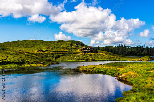 Beautiful shot of a small tranquil pond and river with puffy clouds reflected in water. Scenic green meadows and hills of Connemara, Ireland