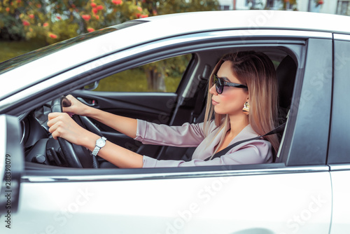 Beautiful business woman lady driving a car in sunglasses, in the summer in the city, a beige formal suit, tanned leather, gold earrings, an expensive branded watch on her hand.
