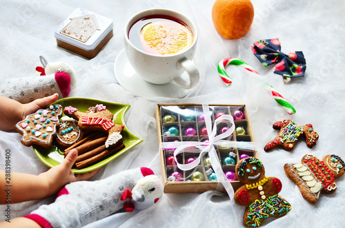 Little child in christmas socks, plate of christmas cookies in hands of baby. Gingerbread man, sweets, cup of tea, box of christmas balls and orange. Table of food and decoration. Close up.
