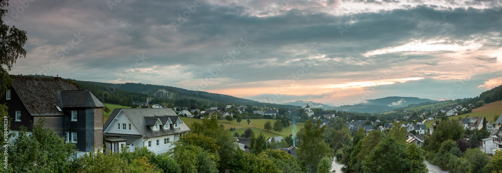 Wide panoramic view over the mountainous village of Grafschaft in the winter sports region of Sauerland, Germany, during sunset on an overcast day with a partly colourful orange lit sky