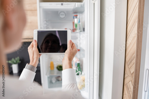 Woman is holding tablet in front of fridge