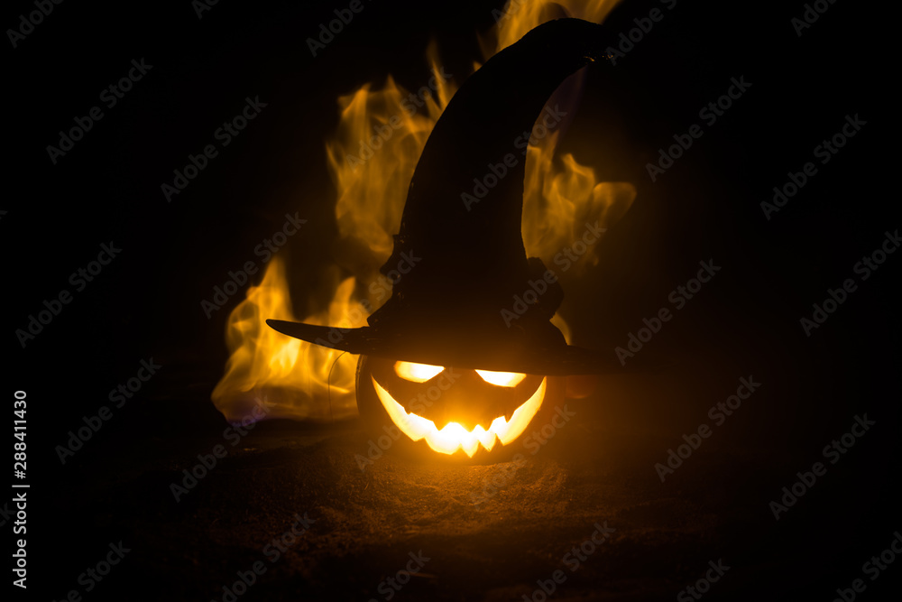 Fototapeta premium Halloween concept. Jack-o-lantern smile and scary eyes for party night. Close up view of scary pumpkin with witch hat on at dark foggy background. Selective focus.