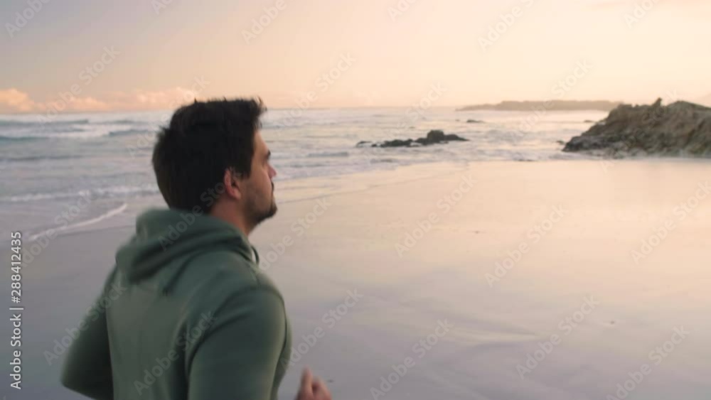 Moving shot of young male jogger in green hoodie jogging along beach in left of frame in late afternoon. Moving shot of young male jogger in green hoodie jogging along beach in left of frame in late afternoon.