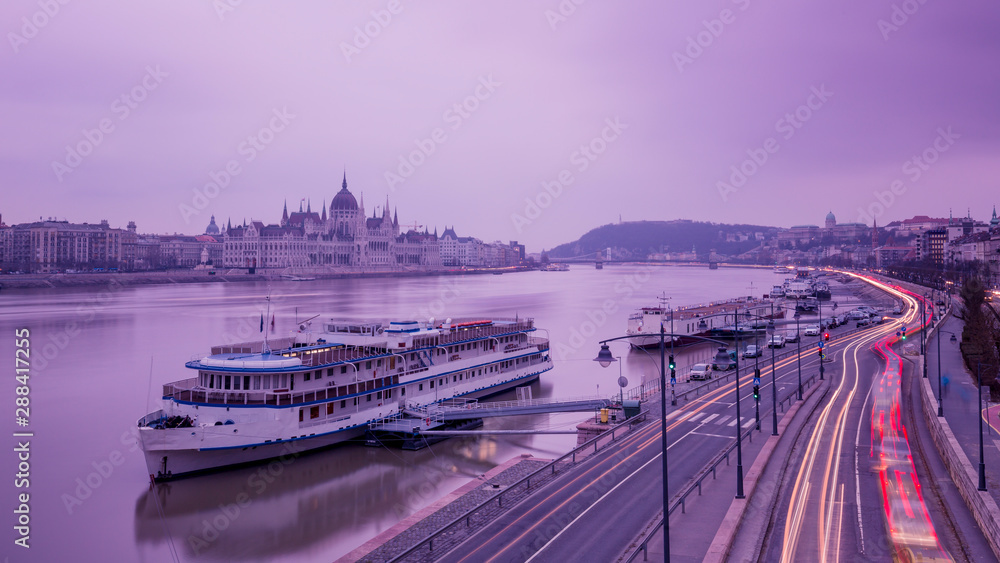 Cityscape of Budapest with parliament and Danube river in overcast sky and cold light. Purple colors, sky reflecting in water and light tracks from moving cars.