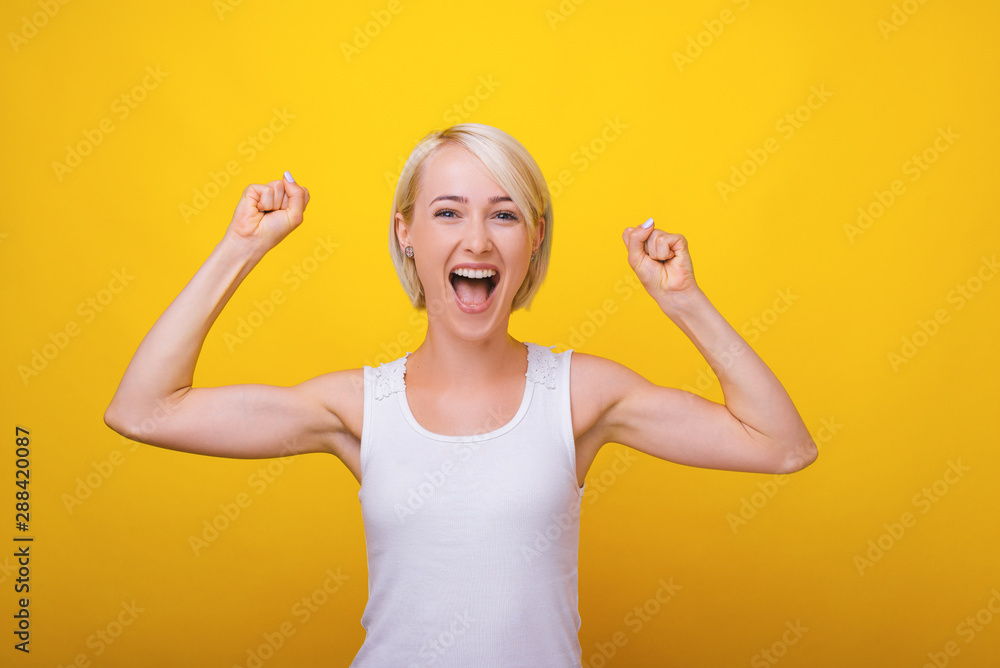 Photo of young blonde woman, celebrating with rised hands and screaming at camera over yellow background Photo of young blonde woman, celebrating with rised hands and screaming at camera over yellow background