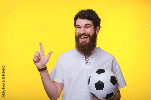 Handsome bearded guyholding soccer ball, while pinting at copy space, yellow background