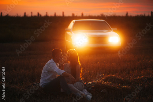 Handsome couple sitting on grass in field, evening on car lights background