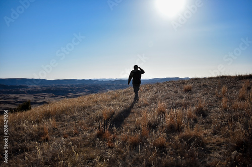 A silhouette of a person walking through rolling hills covered in green and yellow golden grass. The sun in a bright blue sky above beams down on hiker casting a shadow on the ground behind him