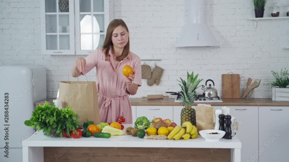 Pretty woman taking out fresh fruit from paper shopping bag at home. Young female holds an orange in the light kitchen. Healthy fruit and vegetables on the table. Pretty woman taking out fresh fruit from paper shopping bag at home. Young female holds an orange in the light kitchen. Healthy fruit and vegetables on the table.