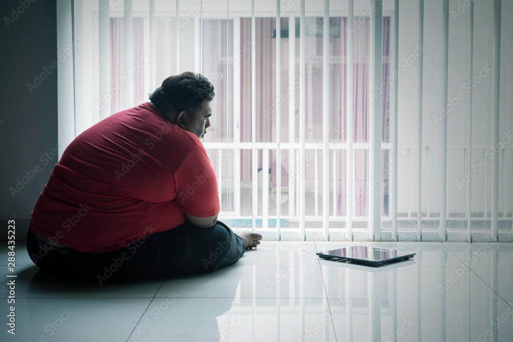 Overweight man sits with a scale near the window Overweight man sits with a scale near the window