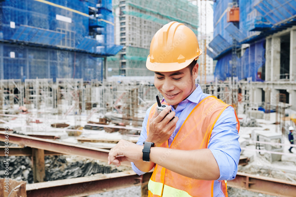 Smiling Asian contractor checking time on his wristwatch when talking to coworkers via walkie-talkie Smiling Asian contractor checking time on his wristwatch when talking to coworkers via walkie-talkie