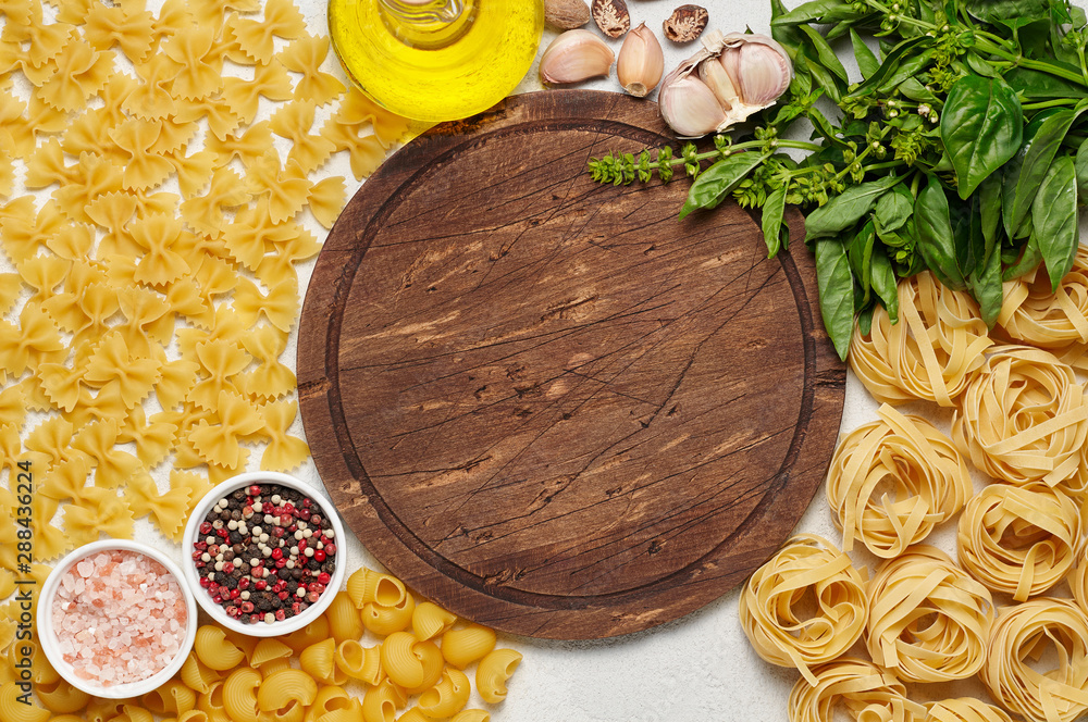 Round rustic wooden cutting board surrounded by different types of Italian pasta, fresh basil, olive oil and garlic