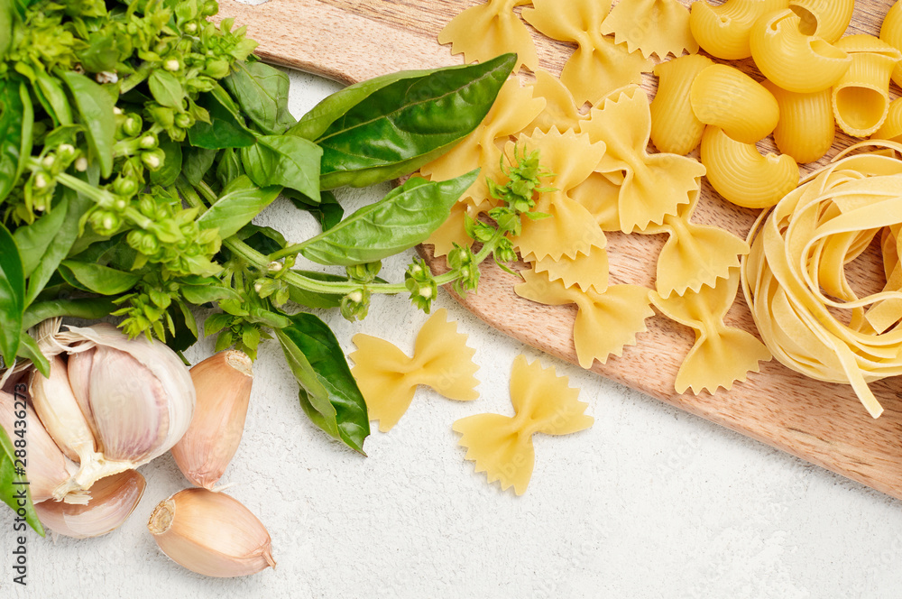 Different types of Italian pasta, fresh basil and garlic on wooden cutting board