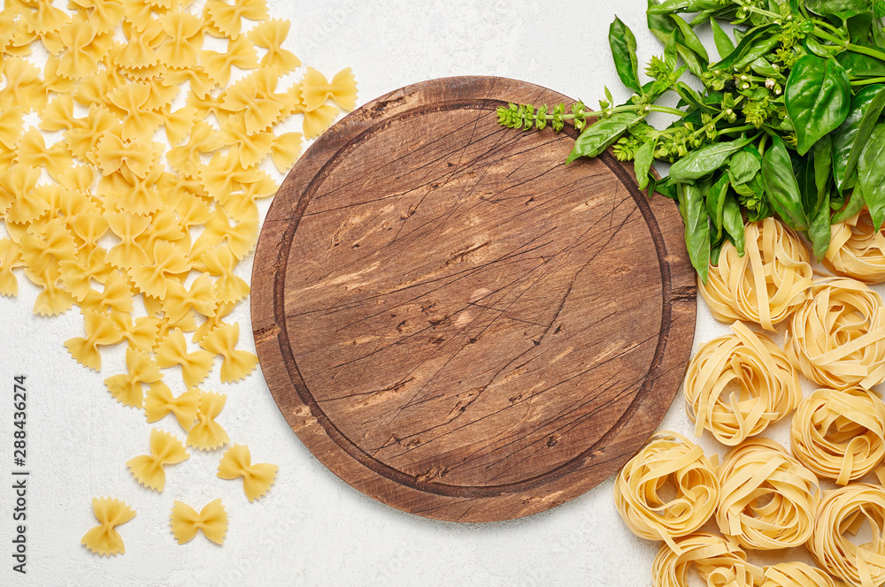 Round rustic wooden cutting board surrounded by different types of Italian pasta and fresh basil