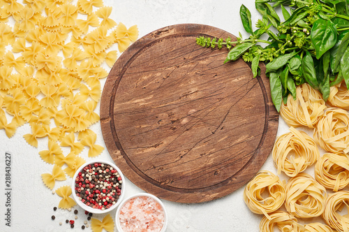 Round rustic wooden cutting board surrounded by different types of Italian pasta