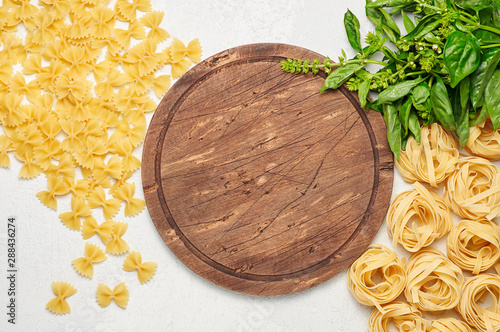 Obraz na płótnie Round rustic wooden cutting board surrounded by different types of Italian pasta and fresh basil w salonie