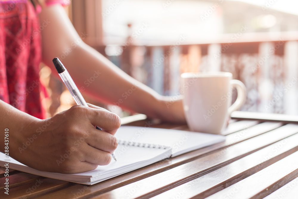 A woman holding a pen is writing on a notebook and a coffee mug on wooden table with warm light A woman holding a pen is writing on a notebook and a coffee mug on wooden table with warm light