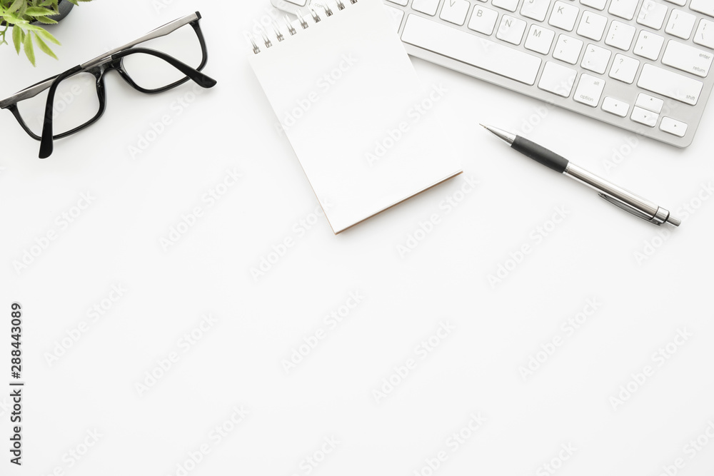 White office desk table with blank small notebook page with pen, computer key board and supplies. Top view with copy space, flat lay. White office desk table with blank small notebook page with pen, computer key board and supplies. Top view with copy space, flat lay.