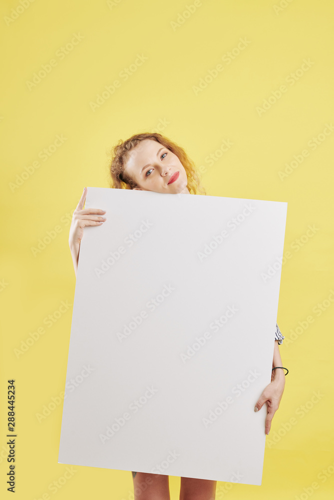 Pensive smiling young woman holding big empty white paper sheet and looking at camera Pensive smiling young woman holding big empty white paper sheet and looking at camera