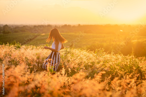 Trendy girl in stylish summer dress feeling free in the field with flowers in sunshine.