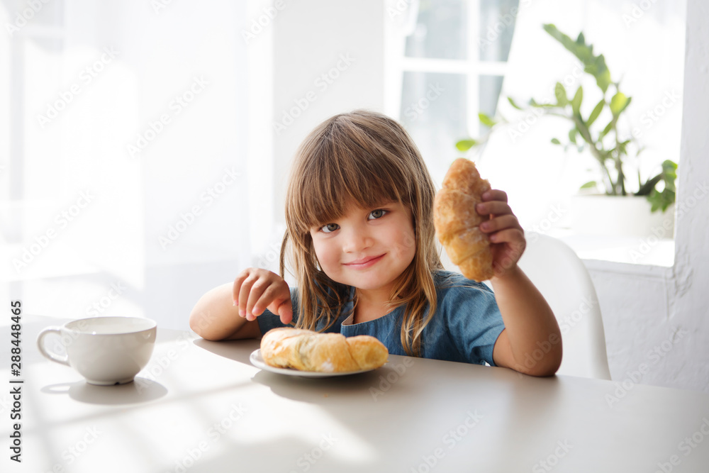 Happy child girll eating croissant and drinking tea in the white kitchen. Happy child girll eating croissant and drinking tea in the white kitchen.