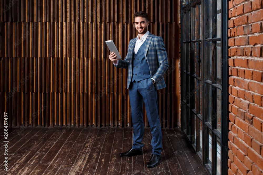 young successful businessman with a beard in a luxurious blue checked suit. Man works on a tablet in a loft office