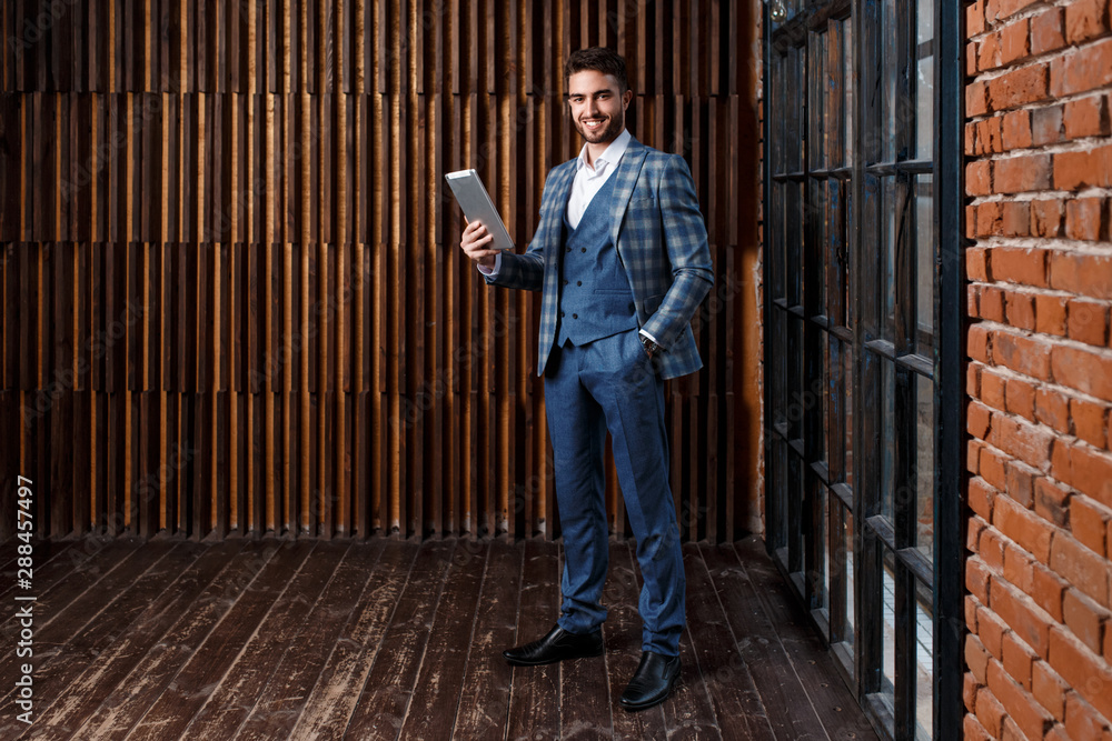 young successful businessman with a beard in a luxurious blue checked suit. Man works on a tablet in a loft office young successful businessman with a beard in a luxurious blue checked suit. Man works on a tablet in a loft office