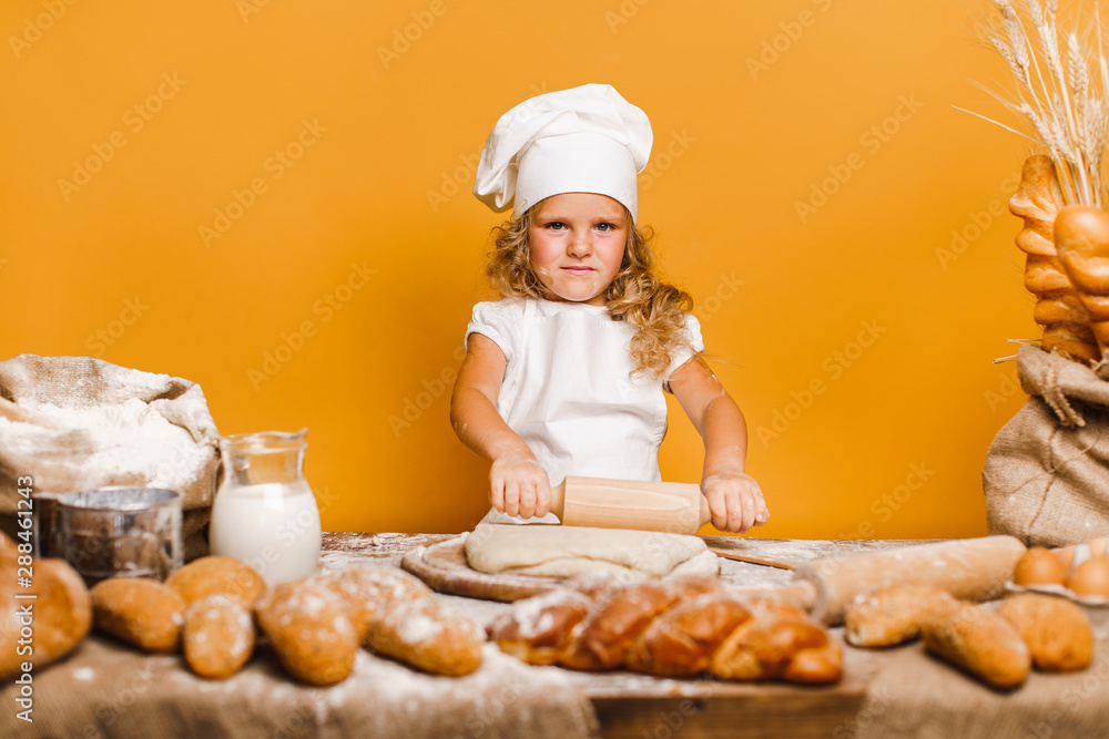 Charming little girl with curly hair in white apron and hat standing at table kneading bread dough and looking at camera. KId in good mood, having fun. Charming little girl with curly hair in white apron and hat standing at table kneading bread dough and looking at camera. KId in good mood, having fun.