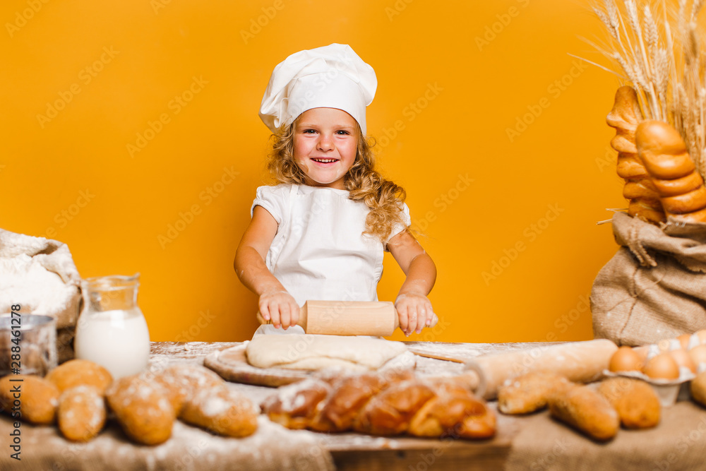 Charming little girl with curly hair in white apron and hat standing at table kneading bread dough and looking at camera. KId in good mood, having fun. Charming little girl with curly hair in white apron and hat standing at table kneading bread dough and looking at camera. KId in good mood, having fun.