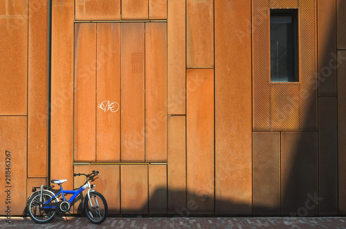 Sunny, rusty facade of an urban building. Modern and geometric corten steel wall covering with a small blue bicycle and a window. Partially shaded wall.