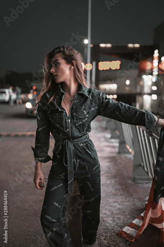 Fashion street portrait of stylish girl in denim overalls on shops background at night. Attractive young woman model walking in city parking in neon light in evening. Urban lifestyle.