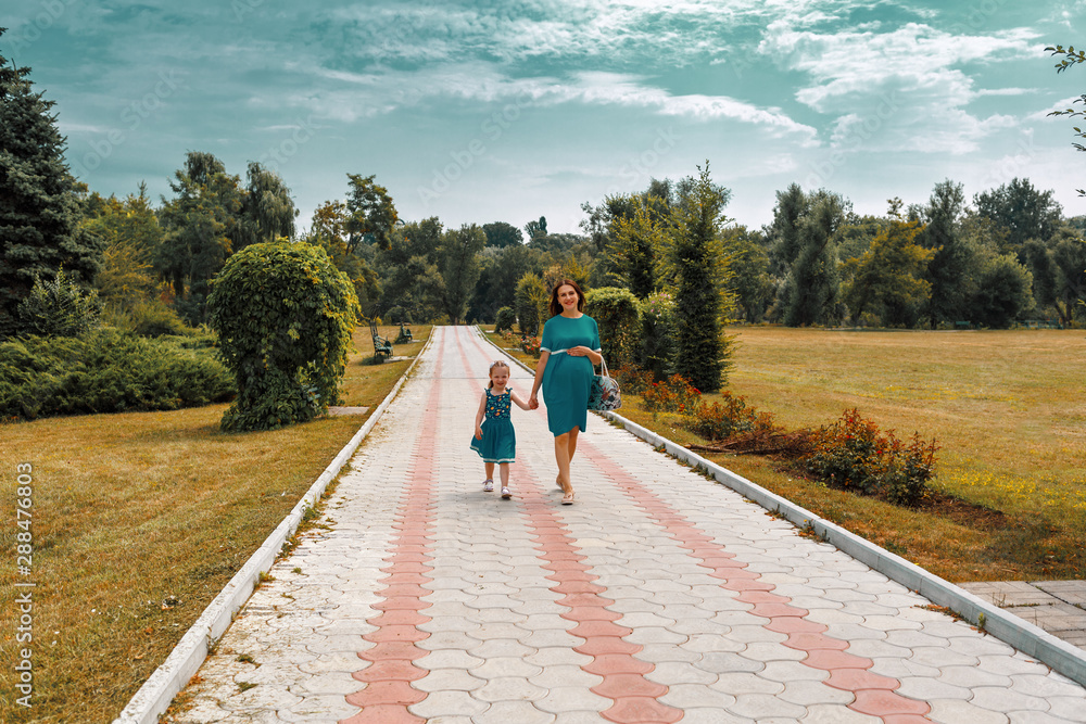 Pregnant woman and a girl holding hands walking in park. Happy family life concept. Bright sunny day and a lot of green plants.