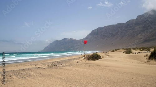 Panoramic view of a beach with a waving red flag and some kite surfers in the background at Caleta de Famara, Lanzarote Island. Slow motion edit photo