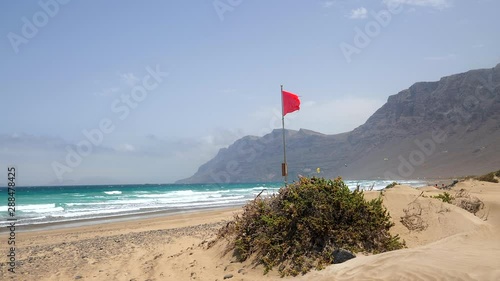 Panoramic view of a beach with a waving red flag and some kite surfers in the background at Caleta de Famara, Lanzarote Island. HD cropped, Slow motion edit photo