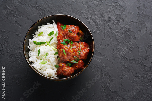 Chicken Manchurian in bowl with basmati rice at black concrete background. Chicken Manchurian is Indian Chinese cuisine dish with Chicken breasts, bell pepper, tomatoes, soy sauce. Copy space