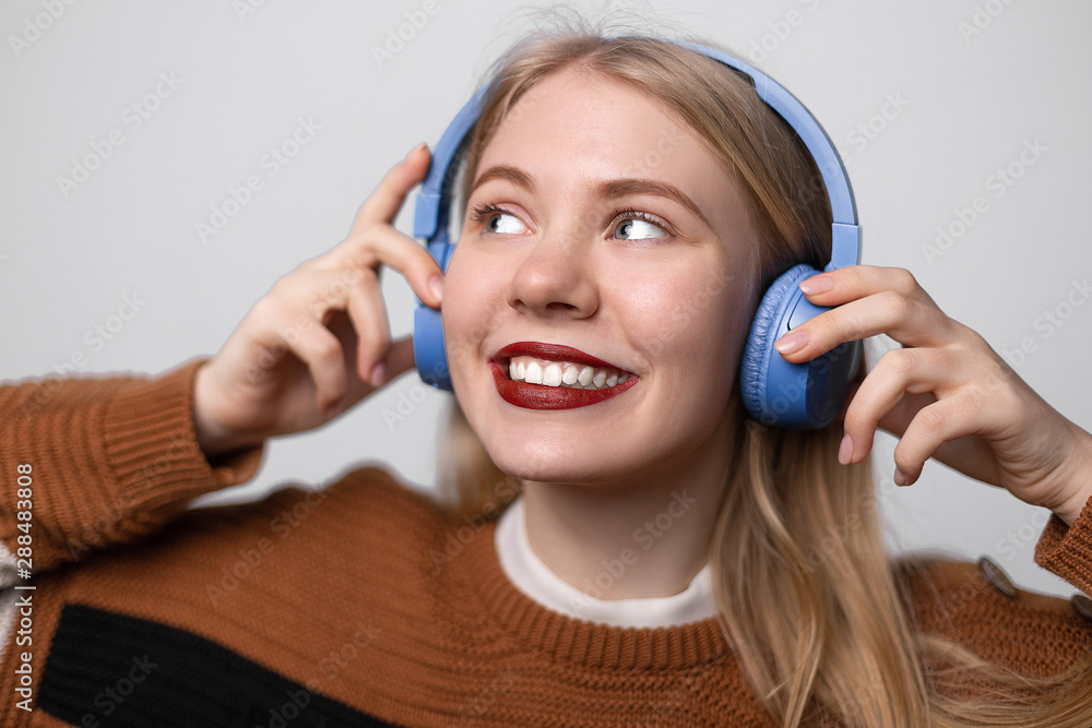 Naklejka premium Happy young blonde female with bright red lips enjoying favorite song while listening it on headphones. Using earphones