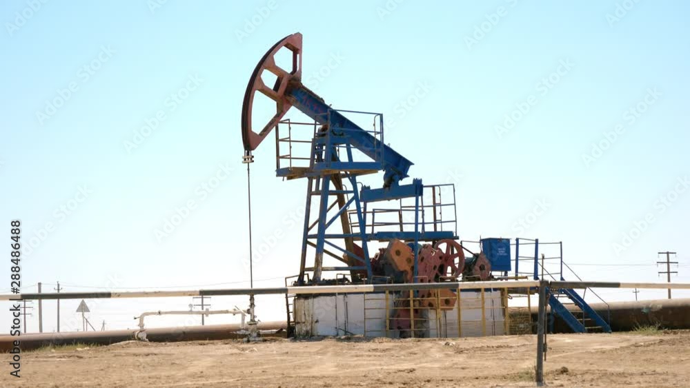CLOSE UP: Industrial oil pump jack working and pumping crude oil for fossil fuel energy with drilling rig in oil field. Nodding donkey pump against the blue sky pumping over the sun in sunny summer CLOSE UP: Industrial oil pump jack working and pumping crude oil for fossil fuel energy with drilling rig in oil field. Nodding donkey pump against the blue sky pumping over the sun in sunny summer