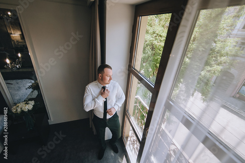 A young groom stands near the window and straightens his tie. The morning of the groom. Wedding portrait of a stylish male businessman. Concept and photography.