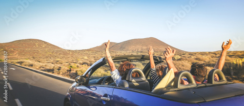 Back view group three euphoric friends in convertible car twisting and waving Two curly girls and charming bearded man on vacation having fun in topless auto Arms up palms open driving in the sunlight photo