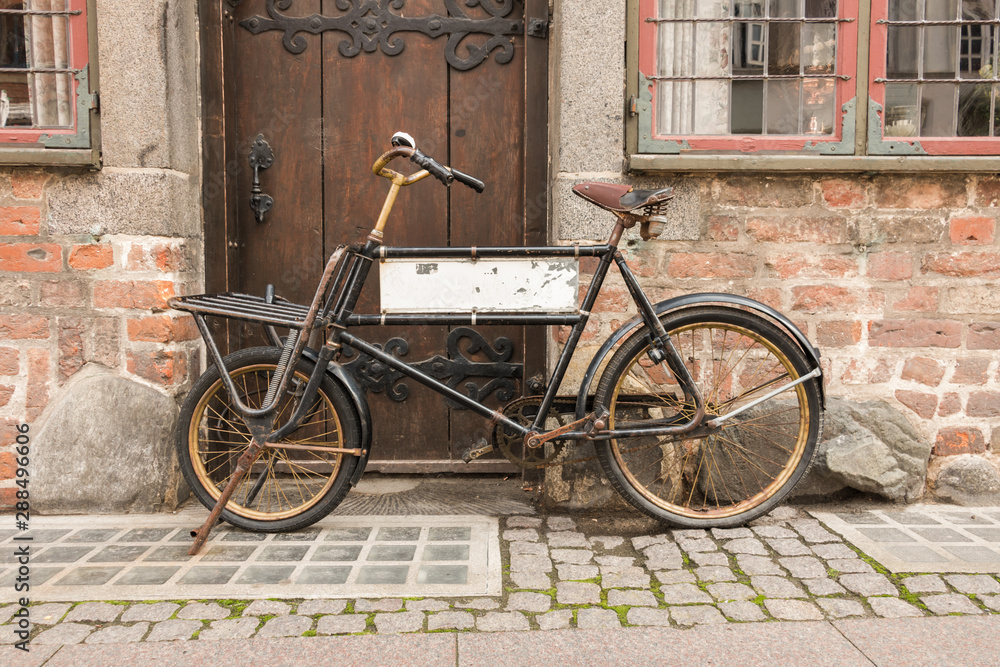 An old bicycle, in front of a brick wall and an old wooden door. The bicycle has a blank sign inside the frame An old bicycle, in front of a brick wall and an old wooden door. The bicycle has a blank sign inside the frame