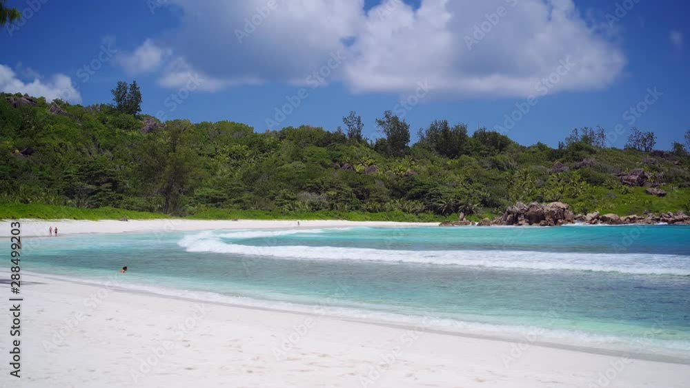 Anse Coco beach on La Digue, Seychelles. Unrecognized tourist swimming in shallow blue ocean lagoon. Calm waves rolling to white sand beach. Blue sky and white clouds above shore. Anse Coco beach on La Digue, Seychelles. Unrecognized tourist swimming in shallow blue ocean lagoon. Calm waves rolling to white sand beach. Blue sky and white clouds above shore.