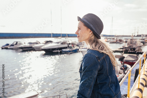 Charming girl tourist enjoys the sunshine standing on the pier and admiring the sea landscape. The concept of vacation at sea in the summer